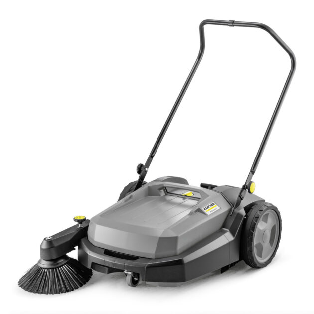 A Sweeper KM 70/20 C with a sturdy metal frame and large black wheels. It features a wide central section with a large front brush to collect debris. The handle is adjustable for ease of use. The sweeper body is mainly gray and black.