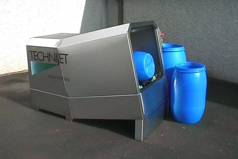An industrial printer labeled "technijet" with a sleek, angular design sits next to two large blue plastic barrels, placed on a rough-textured surface under natural light.