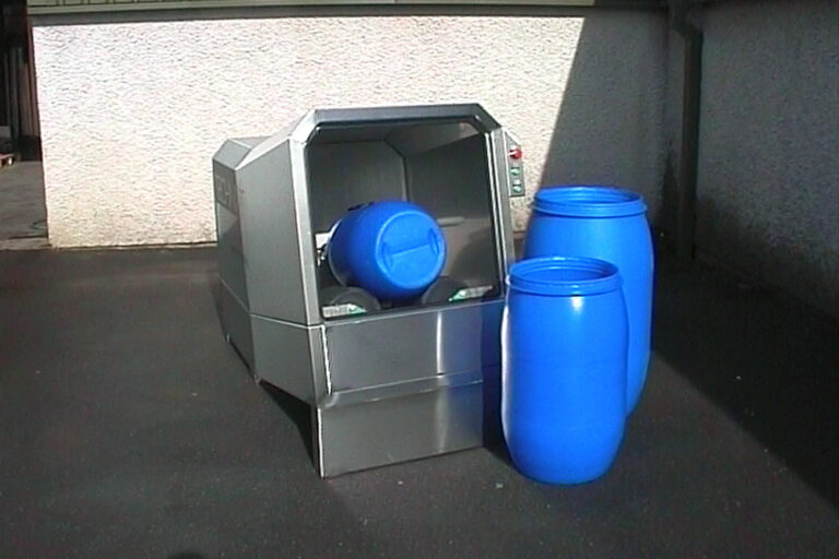 A reverse vending machine for recycling, with a blue plastic barrel being deposited into it, alongside two similar barrels next to the machine on a sunny day.