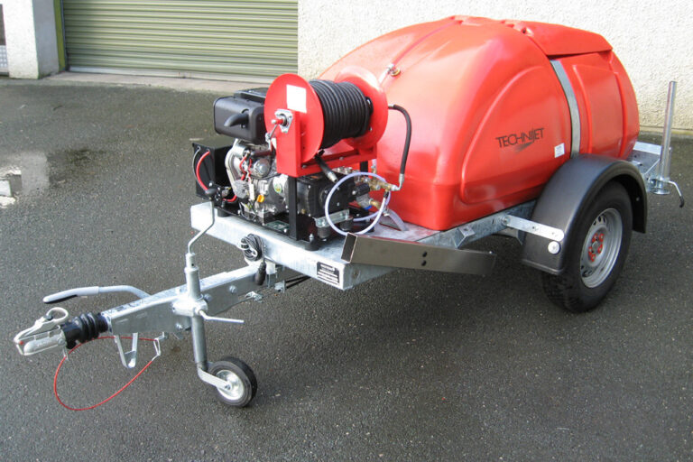 A red industrial pressure washer unit mounted on a small trailer, featuring a large water tank, engine, and a coiled high-pressure hose, parked on a concrete surface near a building.