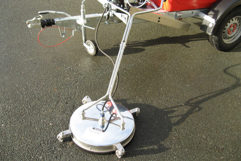A ground-penetrating radar system mounted on a wheeled frame, attached to the back of a small vehicle on a sunny day, with shadows visible on the asphalt surface.