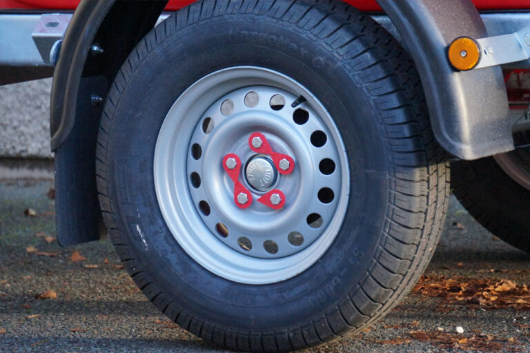 Close-up of a car's wheel with a white steel rim and red lug nuts, parked on a street, showing detailed tire treads and part of the vehicle's body.