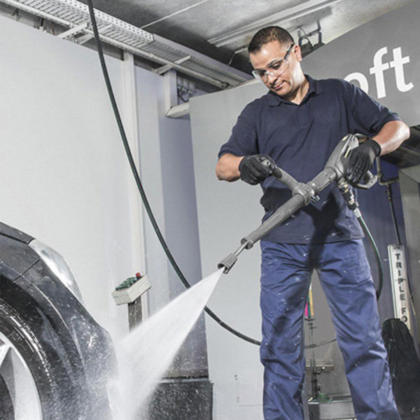 A man in a navy blue work uniform and safety glasses using a high-pressure water spray to clean a large vehicle tire in a professional garage setting.