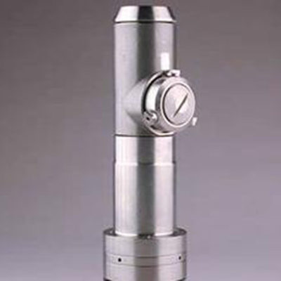 A tall silver cyclindrical microscope with a central viewing piece, standing upright on a gray background.