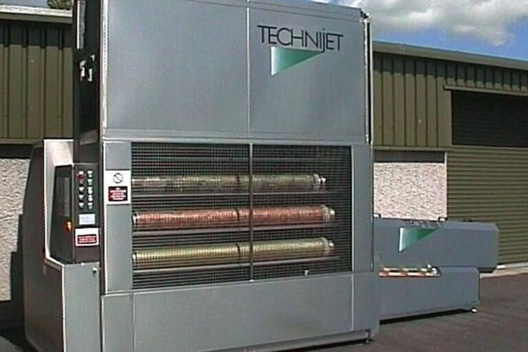 Large industrial printing machine labeled "technijet" with multiple cylindrical rollers visible through a mesh cover, situated outdoors next to storage units.