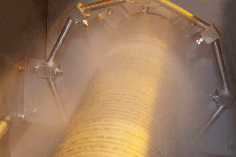 Inside a cylindrical machine, showing a textured metal surface with mist and a warm amber light, surrounded by a metallic structure with bolts and pipes.