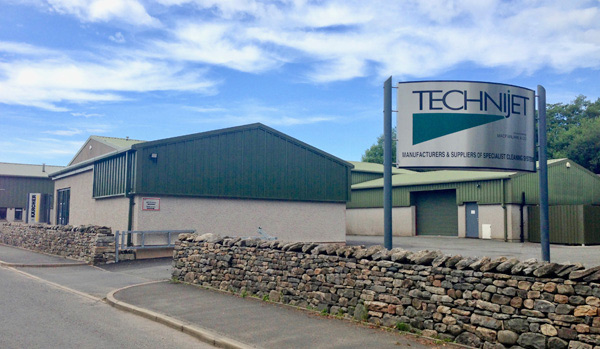 A view of the technijet company facility with a large sign in front, surrounded by a stone wall, under a clear sky. the building has green metal cladding and a flat roof.