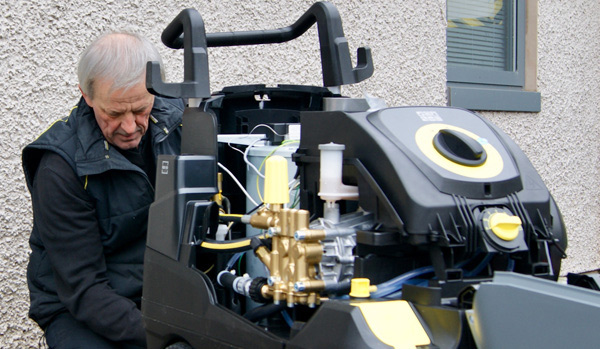 An elderly man examines the internal components of a large, modern machine, concentrating on the intricate arrangement of pipes and gears.