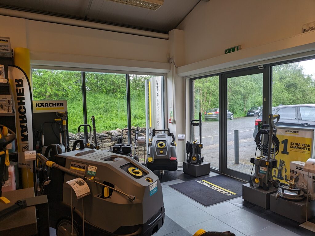 A showroom displaying various Kärcher cleaning equipment, including pressure washers, vacuum cleaners, and other cleaning machines. Large windows let in natural light and provide a view of a parking lot and trees outside. Mats near the entrance feature the Kärcher logo.