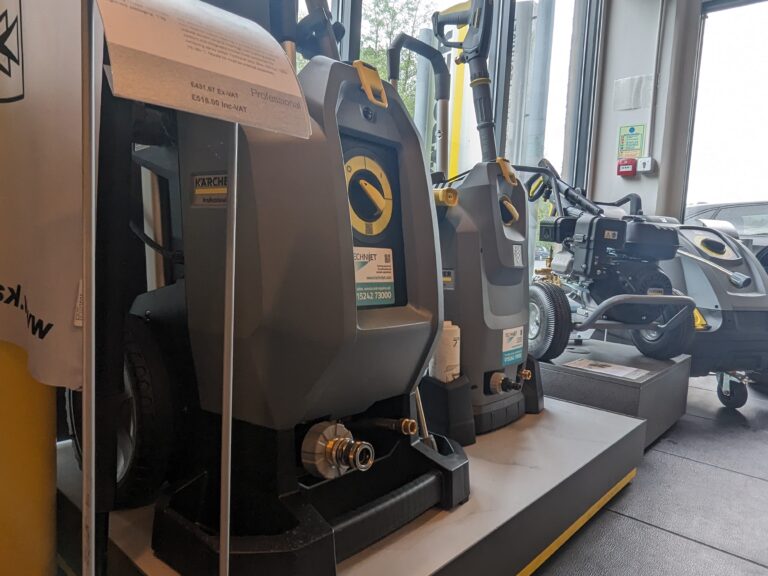 Several Kärcher professional cleaning machines are displayed in a store. The machines are predominantly grey with yellow accents, and are positioned in rows. The background shows a window and a smaller cleaning machine on a raised platform.