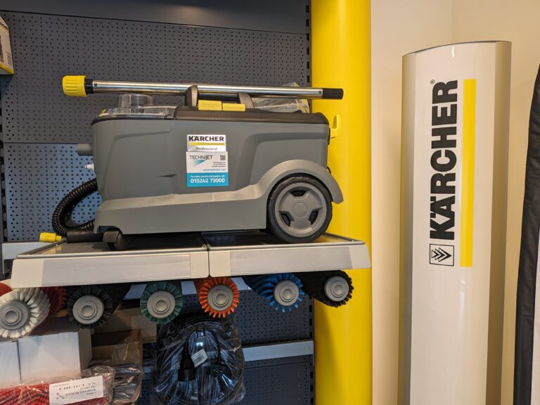 A Karcher cleaning machine is placed on a shelf. It features a gray body with a transparent compartment and a yellow handle on top. Below the shelf are various cleaning brushes. The background includes a gray pegboard and a tall cylindrical Karcher display sign.