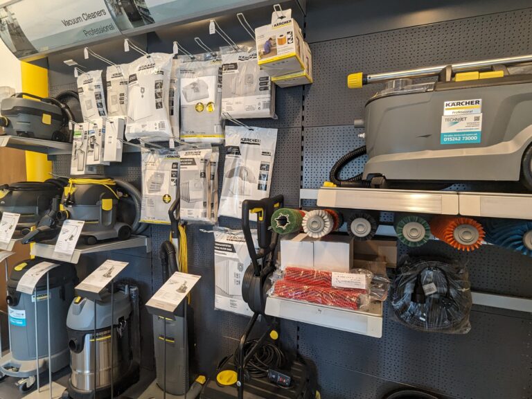 A store display features various Kärcher vacuum cleaners and accessories on shelves and hooks against a pegboard wall. The products include vacuum bags, hoses, cleaning brushes, and industrial vacuum cleaners in different shapes and sizes.