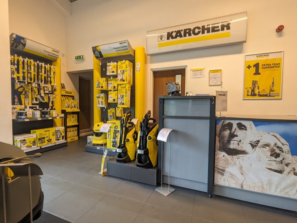 A store interior displaying various Kärcher cleaning products in vibrant yellow and black packaging. The walls have promotional posters and a Mount Rushmore image. A counter stands in the center with brochures and more products neatly arranged on shelves.