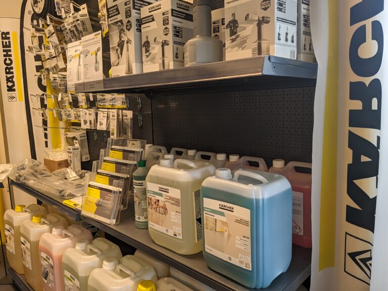 A store shelf displays various Karcher cleaning products, including detergent containers, nozzles, and hoses. The shelf holds multiple bottles of different cleaning solutions alongside boxed equipment. The left and right sides of the shelf feature Karcher branding.