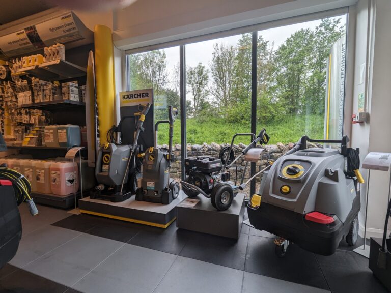 A display of Kärcher cleaning equipment in a store showroom near large windows. Various high-pressure washers and cleaning supplies are neatly arranged on platforms, with a green, tree-lined view outside the windows. Shelves on the left hold additional products.