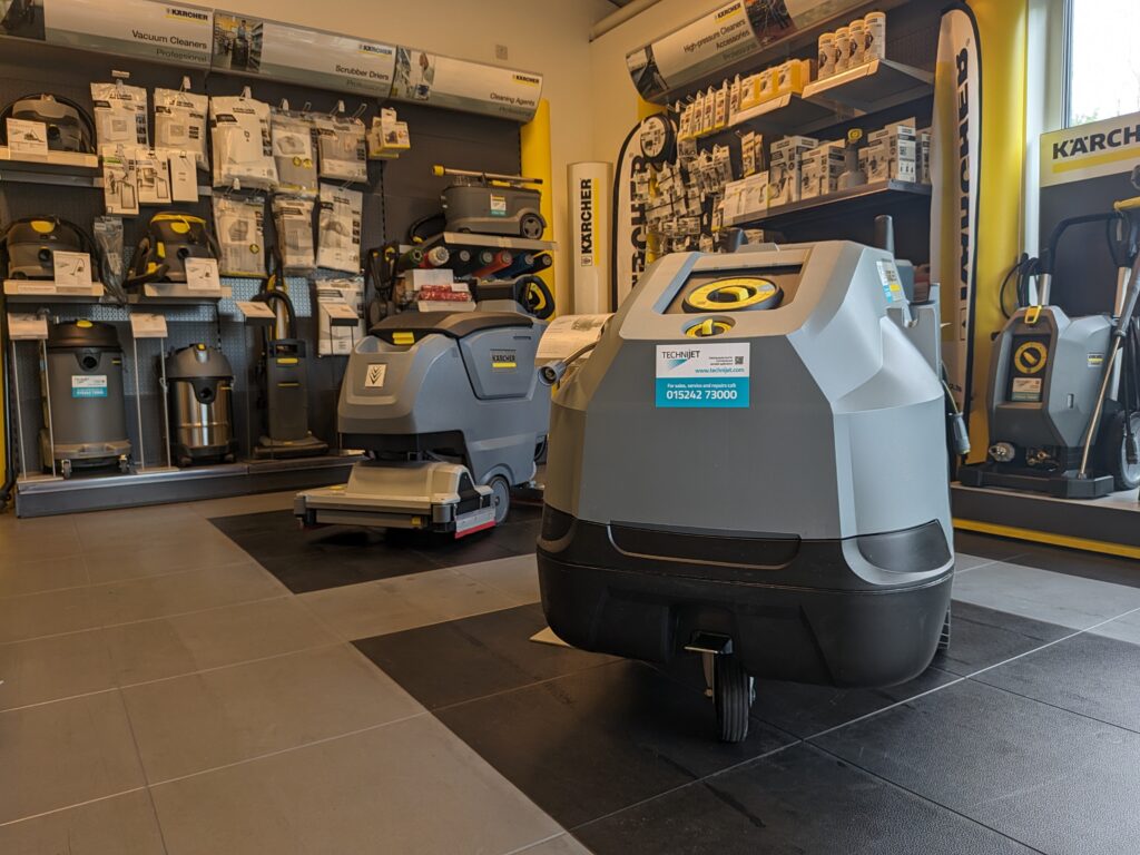 A showroom displays several floor cleaning machines and equipment. The main focus is on two large grey and black machines in the center, with shelves in the background stocked with various cleaning accessories and parts. Karcher brand products are visible throughout.