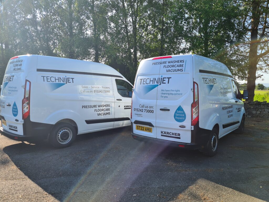 Two white vans parked outdoors, both branded with "TECHNIJET" and other contact details. The vans promote pressure washers, floor care, and vacuum services. The background includes trees and a stone wall under a sunny sky.